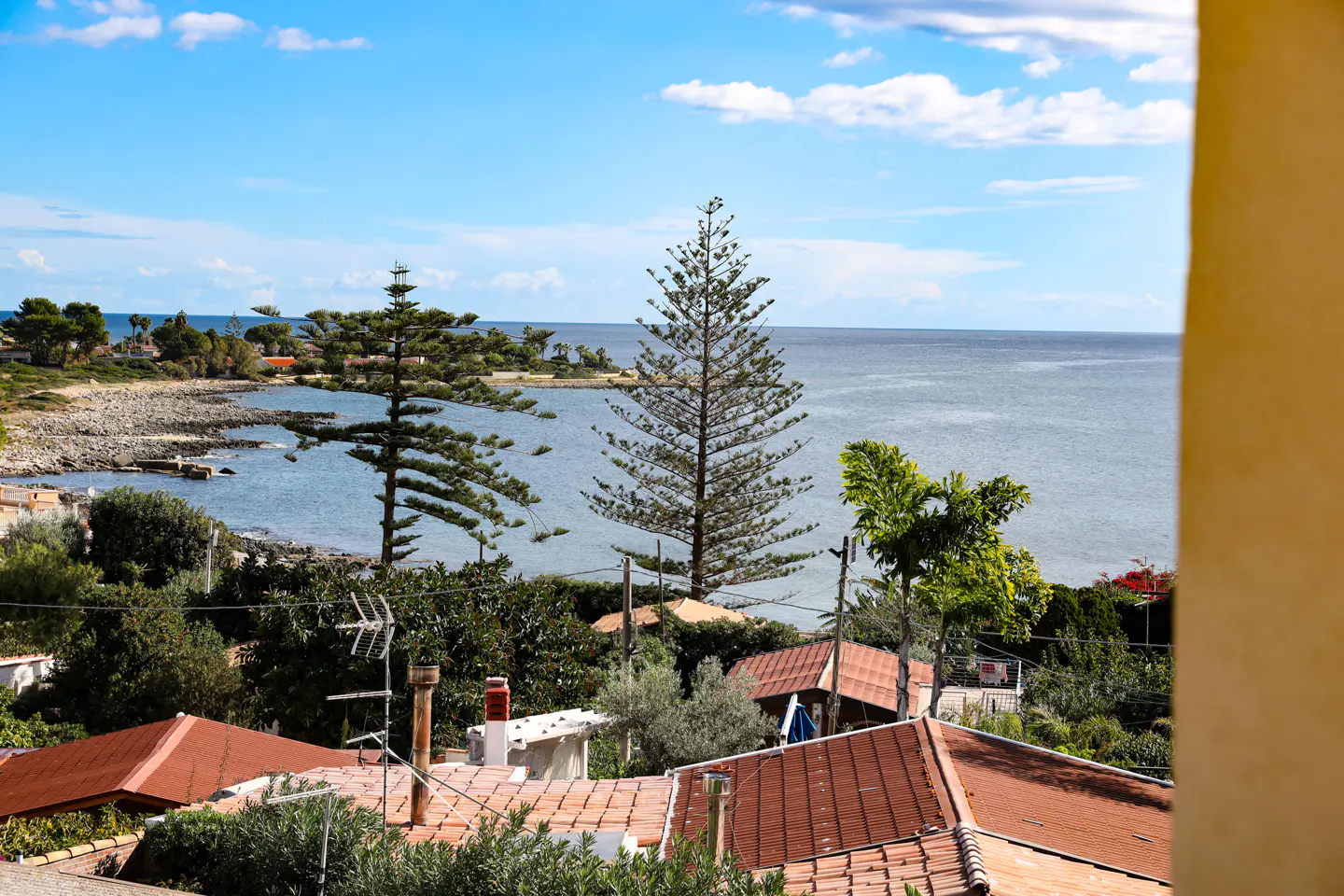 Scenic view of the ocean and coastline, with trees, rooftops, and a blue sky with clouds.