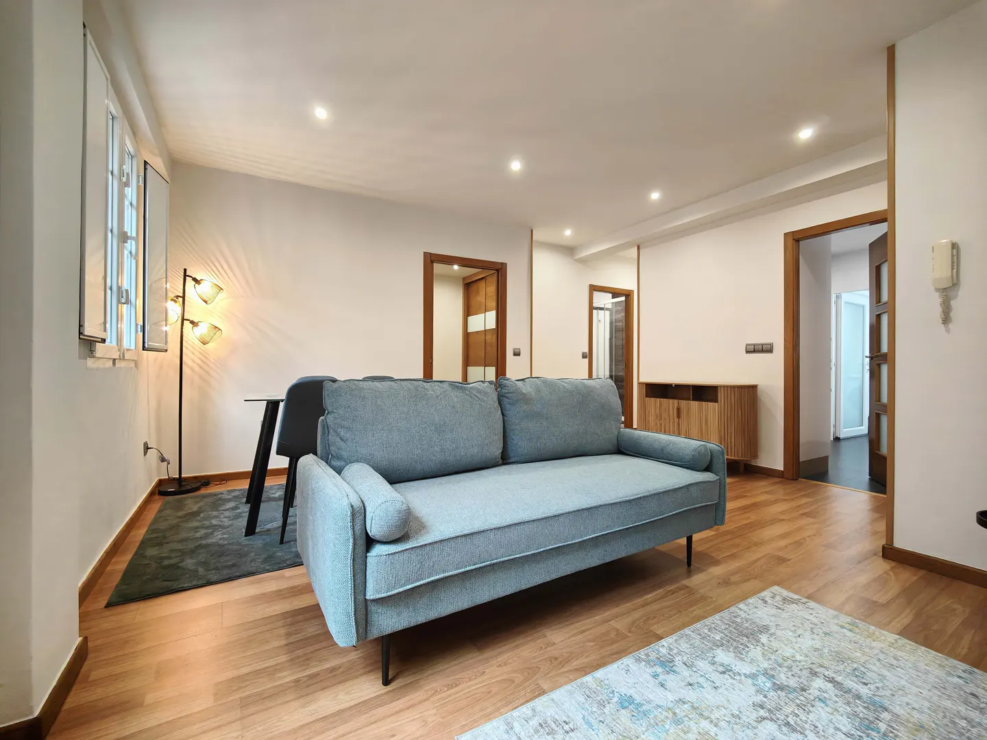Bright living room with a blue sofa, wood floors, and white walls. A dining table, lamp, and doors are visible.