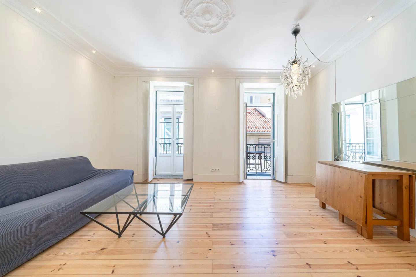 Bright living room with light wood floors, a grey sofa, glass coffee table, crystal chandelier, and two balcony doors.
