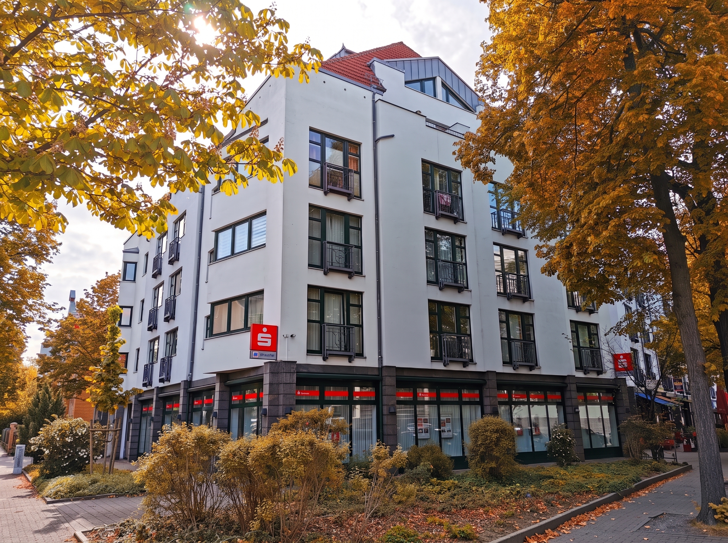 A white multi-story building with a red roof and ground-floor bank branch, framed by autumn trees on a sunny day. [AI-generated text]