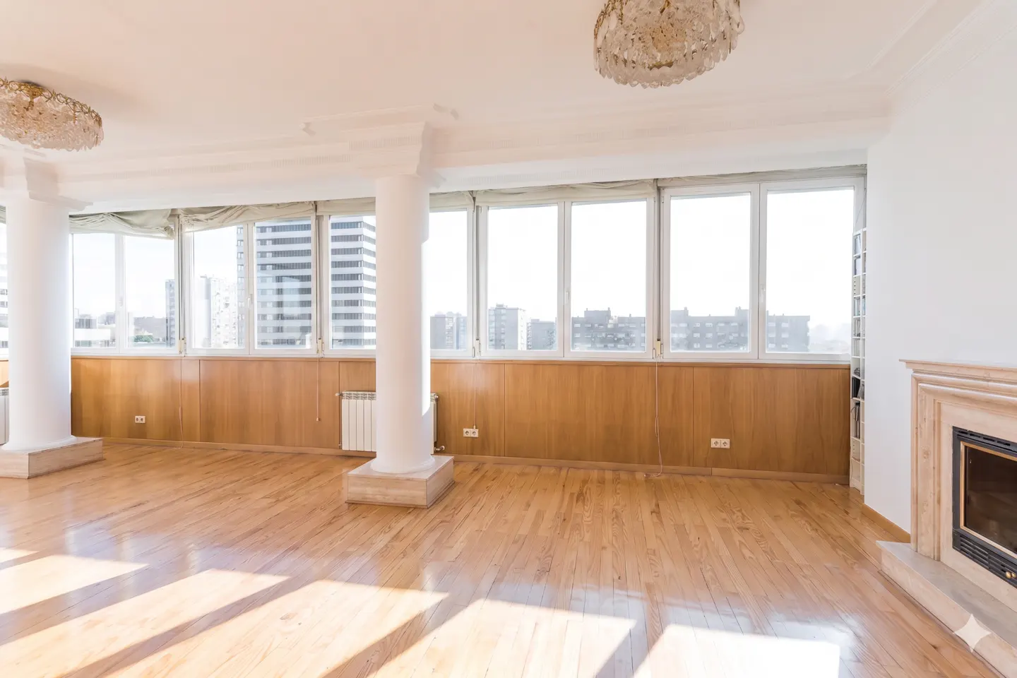Bright, empty room with wood floors, white walls, and large windows with city views. Columns and a fireplace add architectural detail. Chandeliers hang from the ceiling.