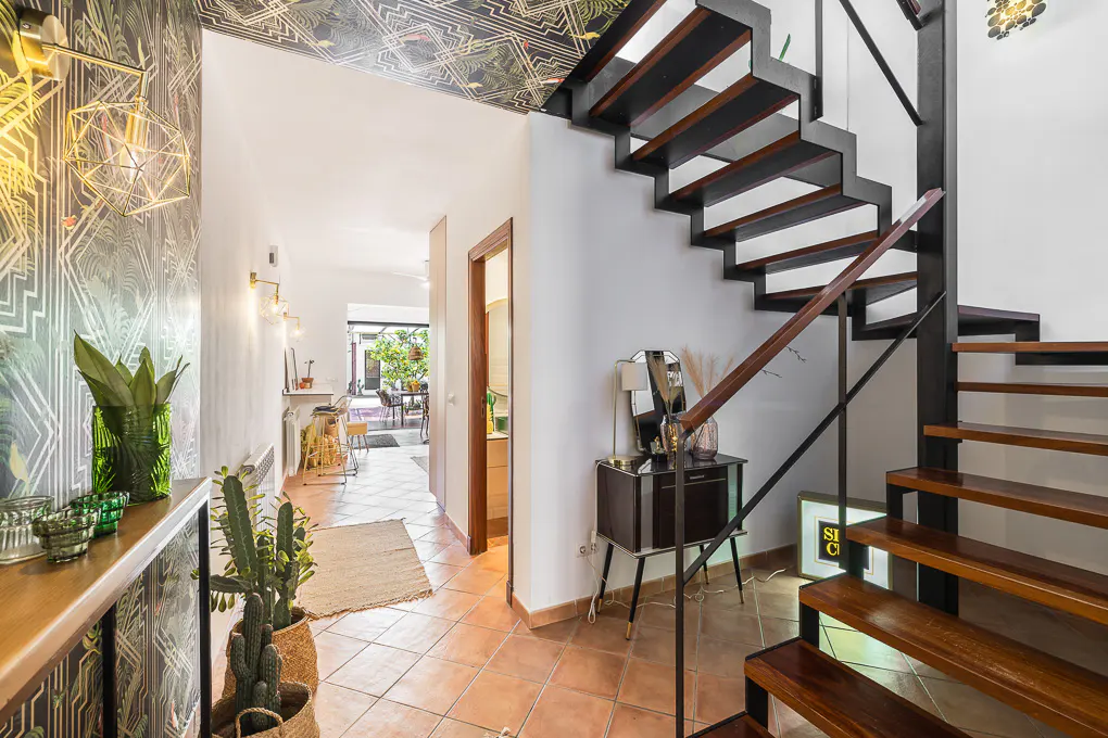 Hallway with terracotta tile floor, black and wood stairs, patterned wallpaper, and a view to an outdoor dining area.
