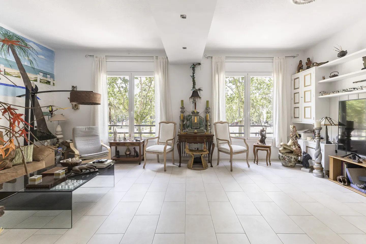 Bright living room with white tile floor, eclectic decor, and large windows with sheer curtains. A glass coffee table sits in the foreground.