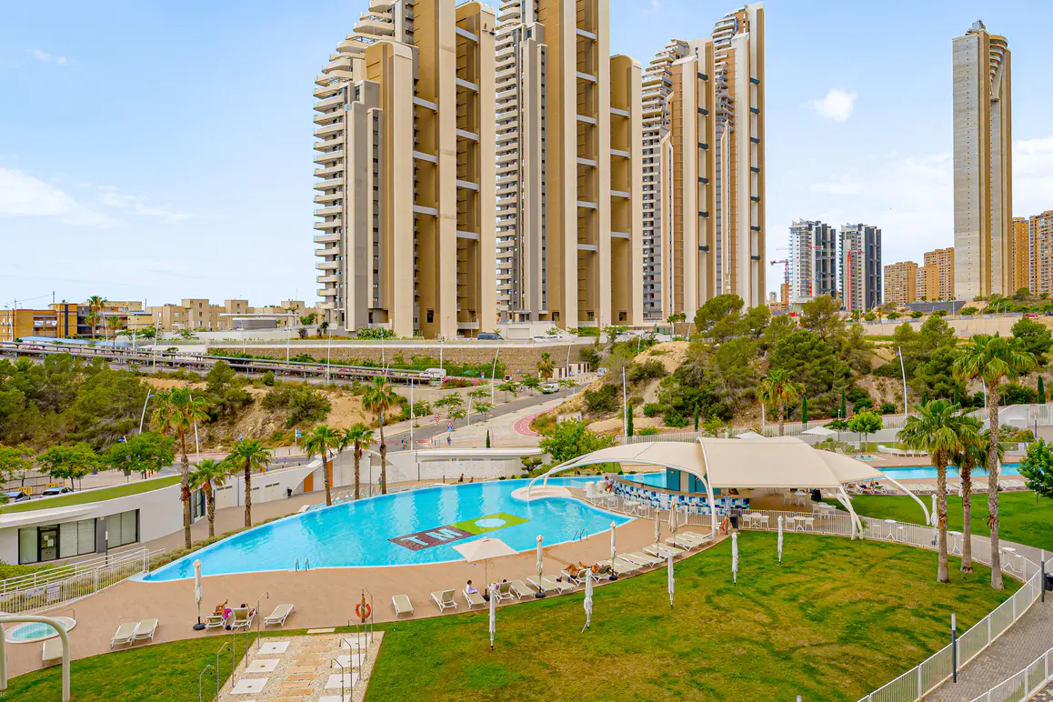 A large turquoise swimming pool with lounge chairs and a shaded bar area, set against a backdrop of modern high-rise towers.