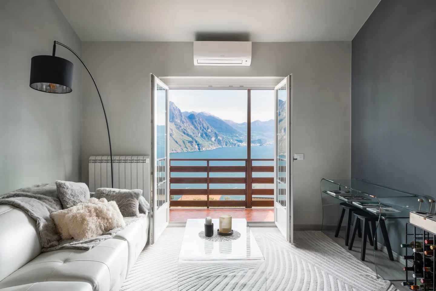 Bright living room with white sofa, glass table, and open doors to a balcony with a lake and mountain view.