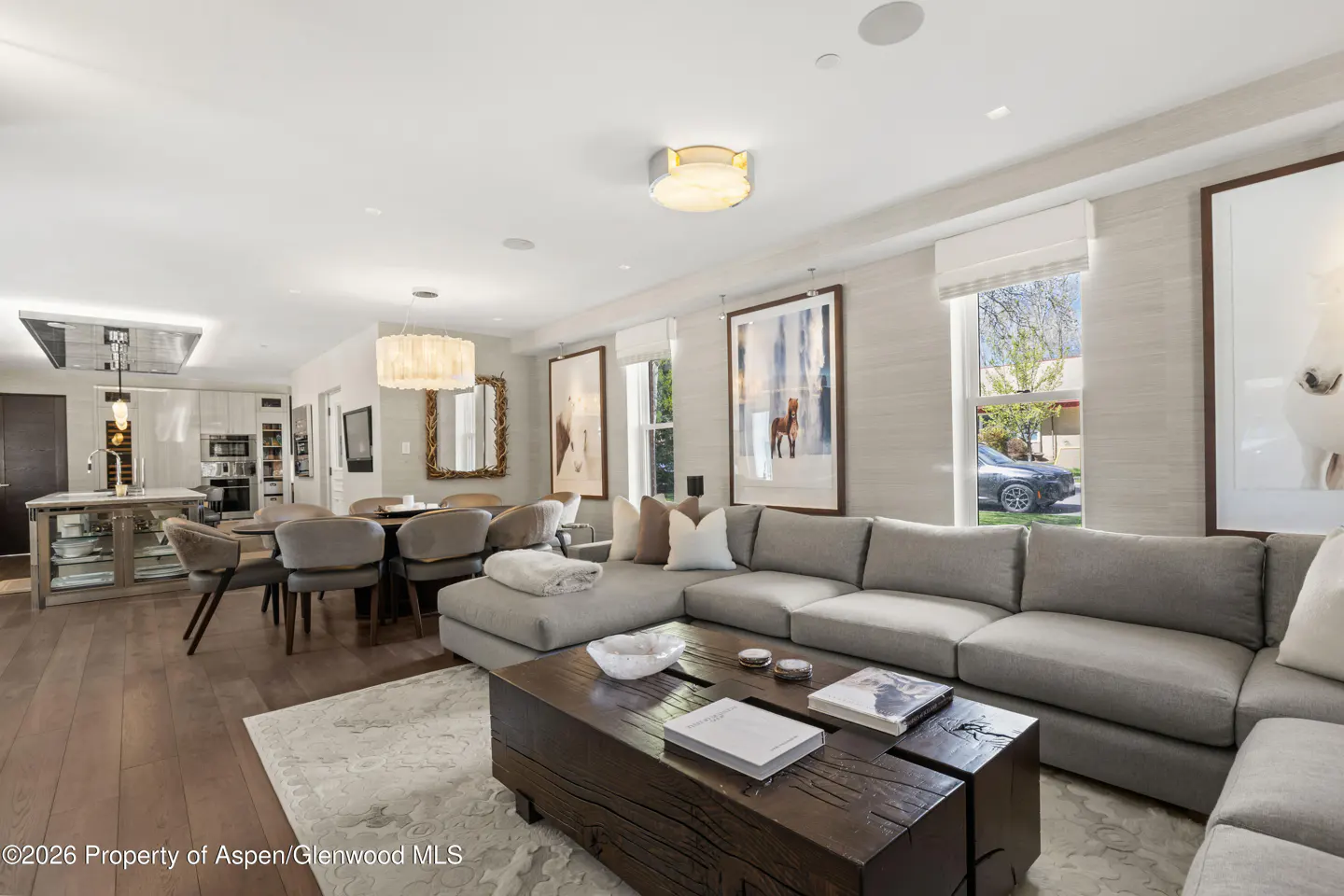 Open-concept living space with a gray sectional sofa, wood coffee table, dining area, and kitchen. Artwork of horses adorns the walls.