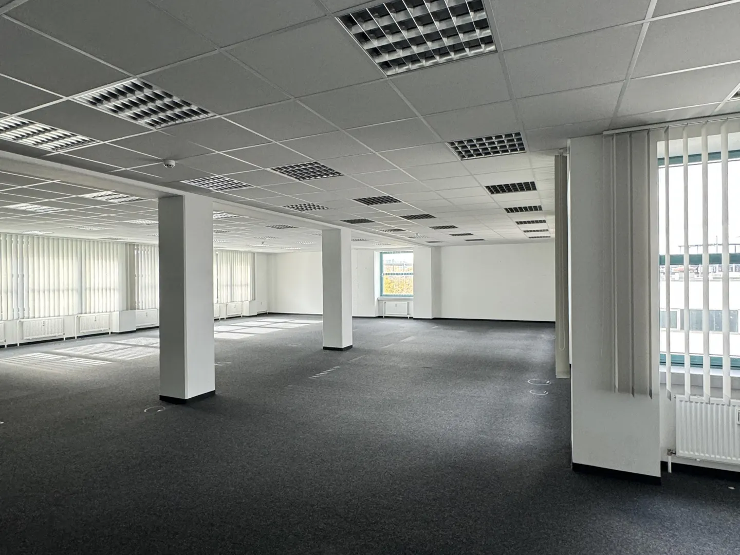 Empty office space with gray carpet, white walls, and support columns. Windows with blinds line the walls, and a grid of lights covers the ceiling.