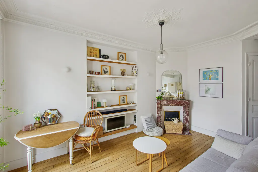 Bright living room with white walls, wood floors, and built-in shelves. A marble fireplace, round tables, and a gray sofa add to the cozy atmosphere.