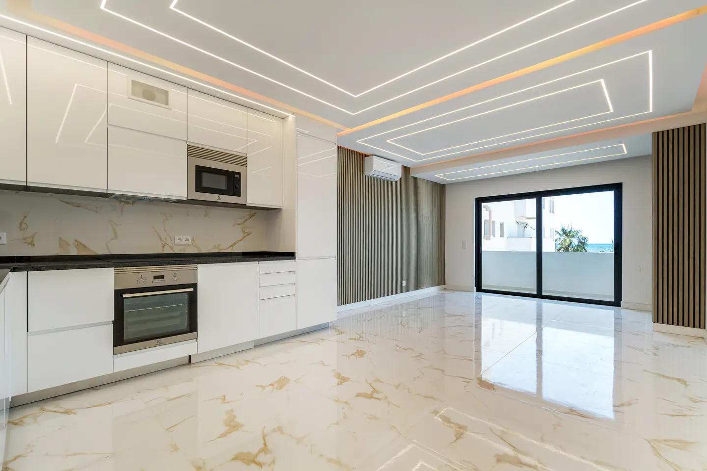 Modern kitchen with white cabinets, marble-look floors, and recessed ceiling lighting, opening to a balcony with sea view.
