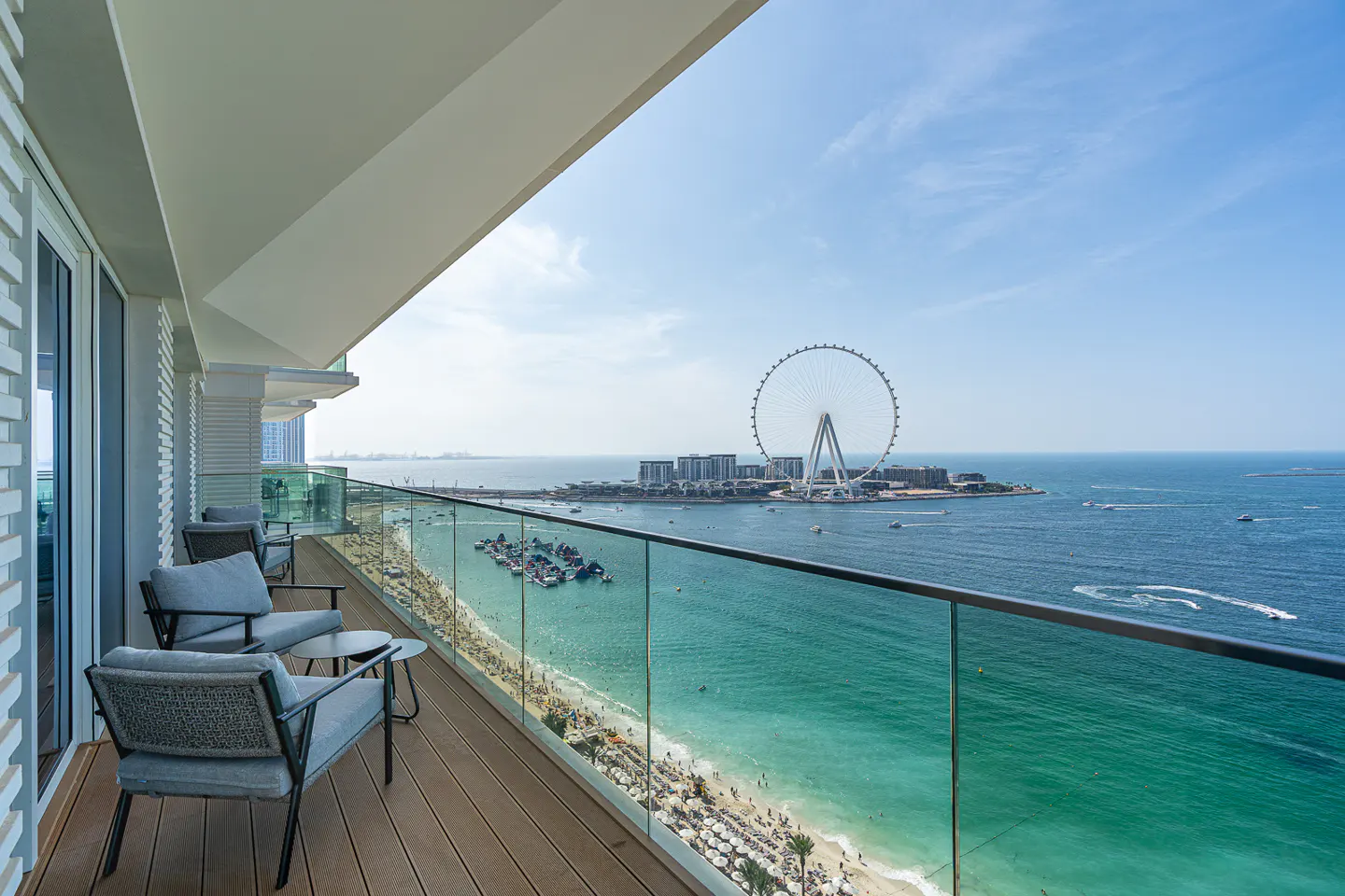 Balcony view of Dubai Eye, beach, and ocean. Gray chairs sit on a wood deck with a glass railing. Blue sky above.