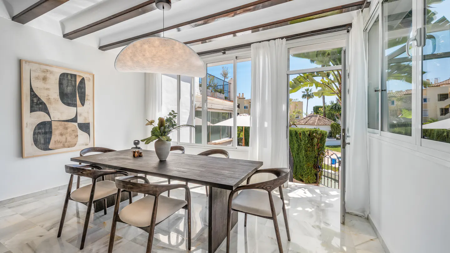 Bright dining room with a dark wood table, six chairs, and marble floors. Large windows overlook a lush garden. Modern art hangs on the wall.
