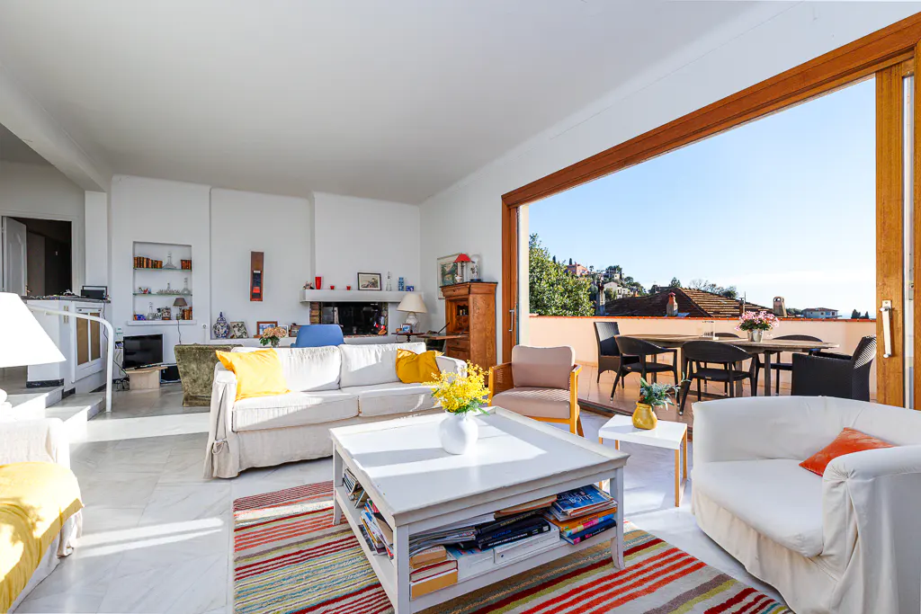 Bright living room with white sofas, a colorful rug, and a view of a terrace with outdoor furniture.