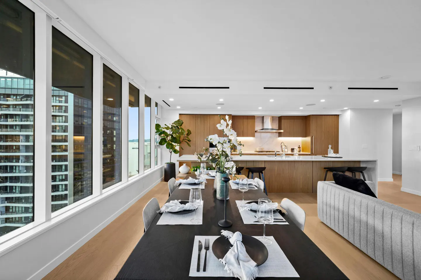 Bright, modern condo interior with a long dining table set for a meal, open kitchen, and city views through large windows.