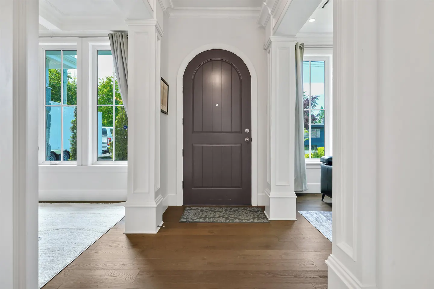 A foyer with dark wood floors, white walls, and a dark brown arched front door. Windows on either side of the door let in natural light.