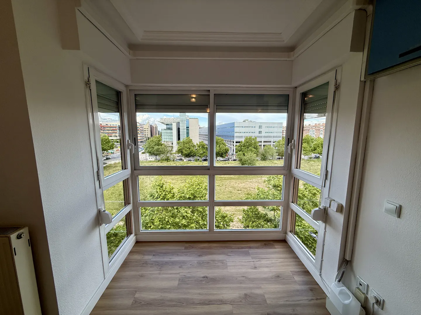 A bright sunroom with white walls and wood floors has large windows overlooking a green field and city buildings.