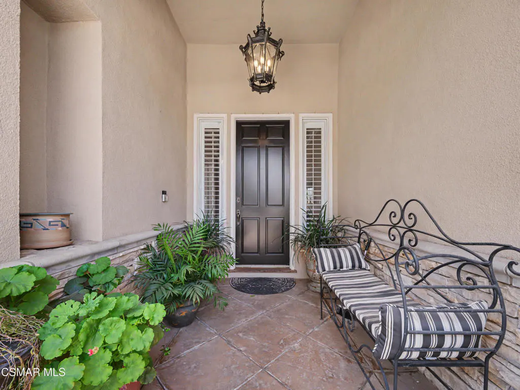 Covered entryway with a black front door, hanging lantern, potted plants, and a striped metal bench on tiled flooring.