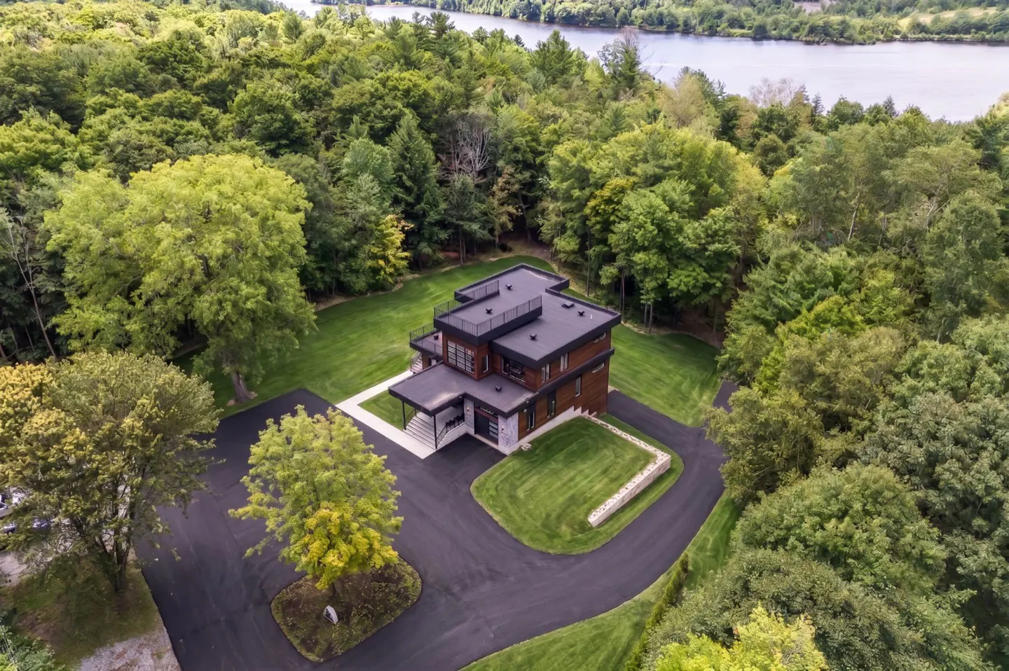 Aerial view of a modern multi-level home with a flat roof, surrounded by lush green forest and a river in the background.