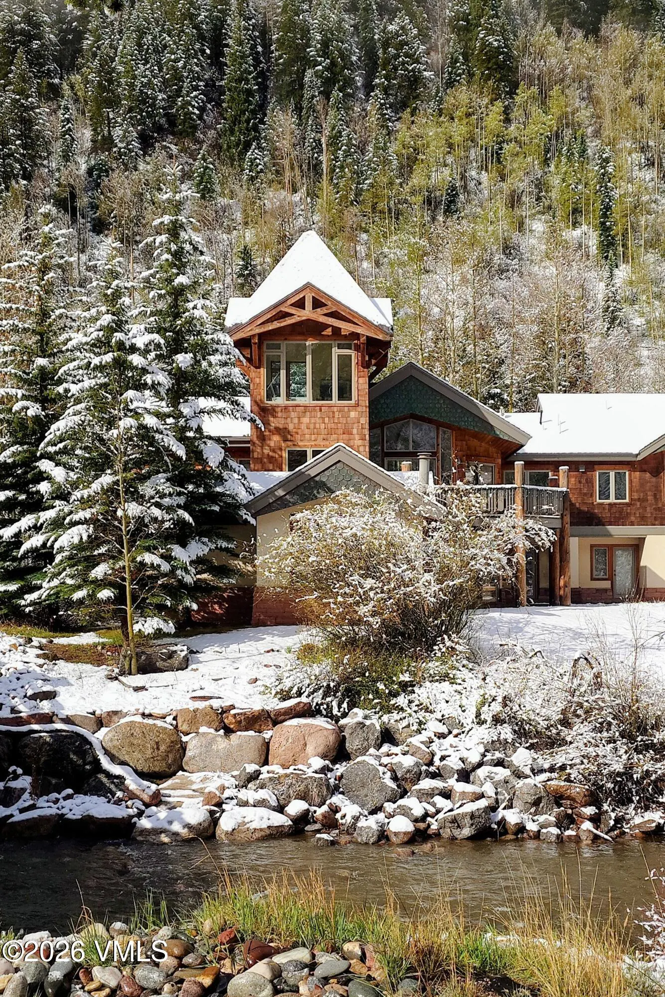 A multi-level wood home sits near a stream, surrounded by snow-covered trees and rocks.