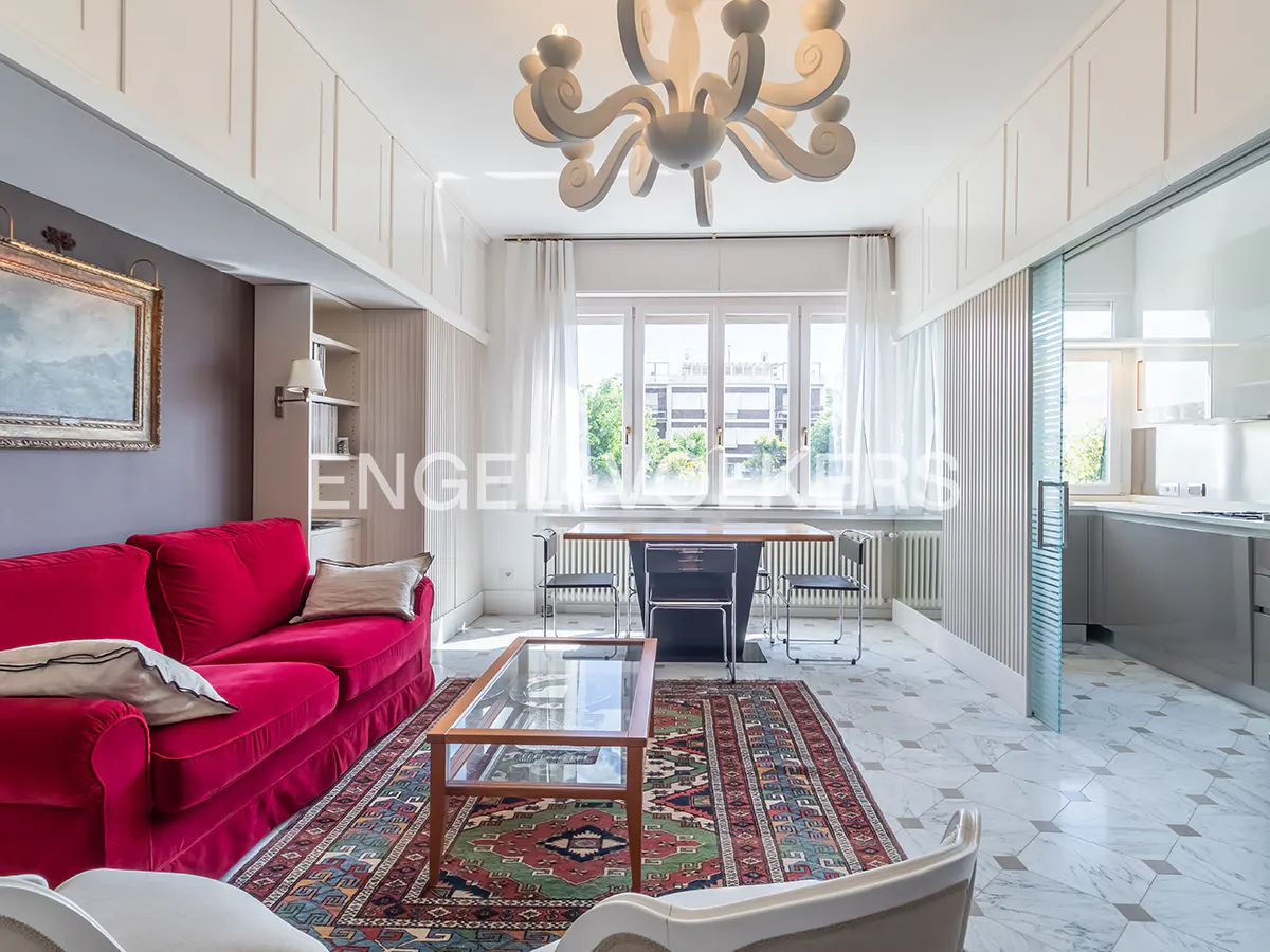 Bright living room with red sofa, patterned rug, glass coffee table, and dining area with large windows. A modern chandelier hangs from the ceiling.