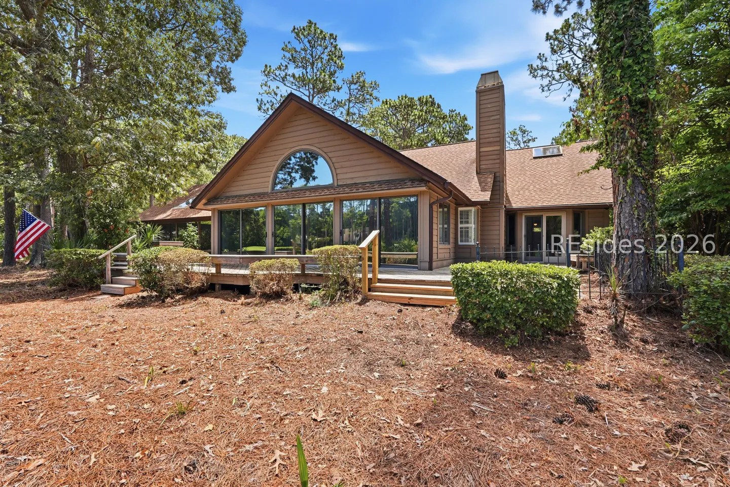 Brown house with a large deck, glass sliding doors, and a chimney, set in a wooded yard with an American flag.