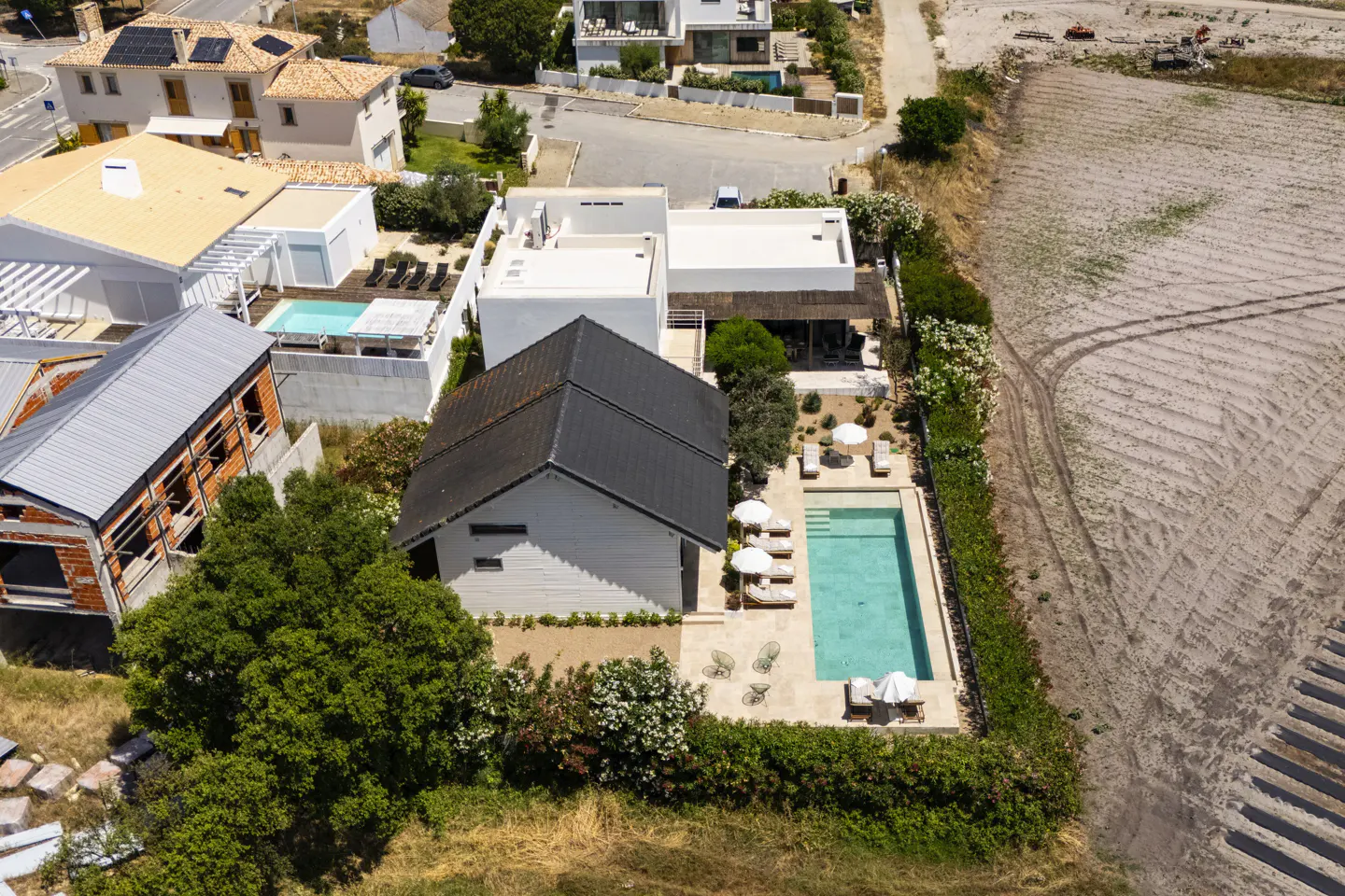 Aerial view of a modern white villa with a dark roof, private swimming pool, and sun loungers next to an empty dirt lot.