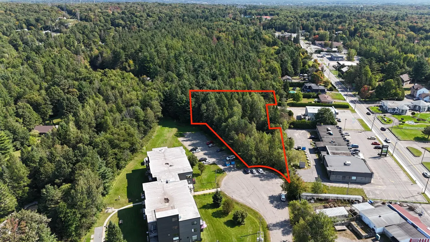 Aerial view of a wooded lot outlined in red, adjacent to a parking lot and buildings. Trees and a road are in the background.