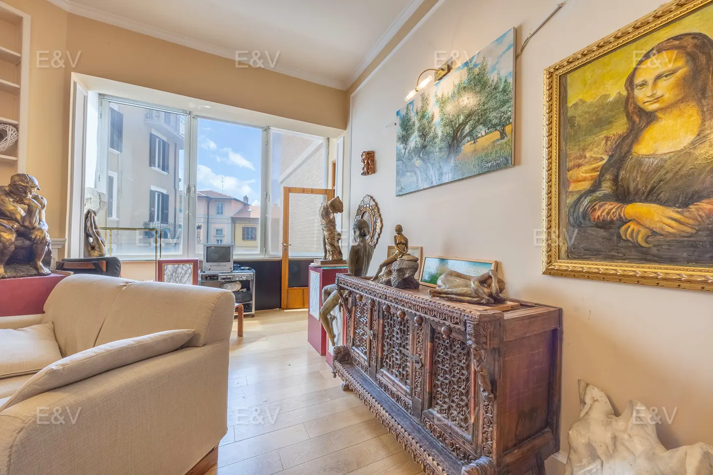 Living room with beige sofa, carved wooden cabinet, and art including a Mona Lisa painting. Outside the window is a city view.