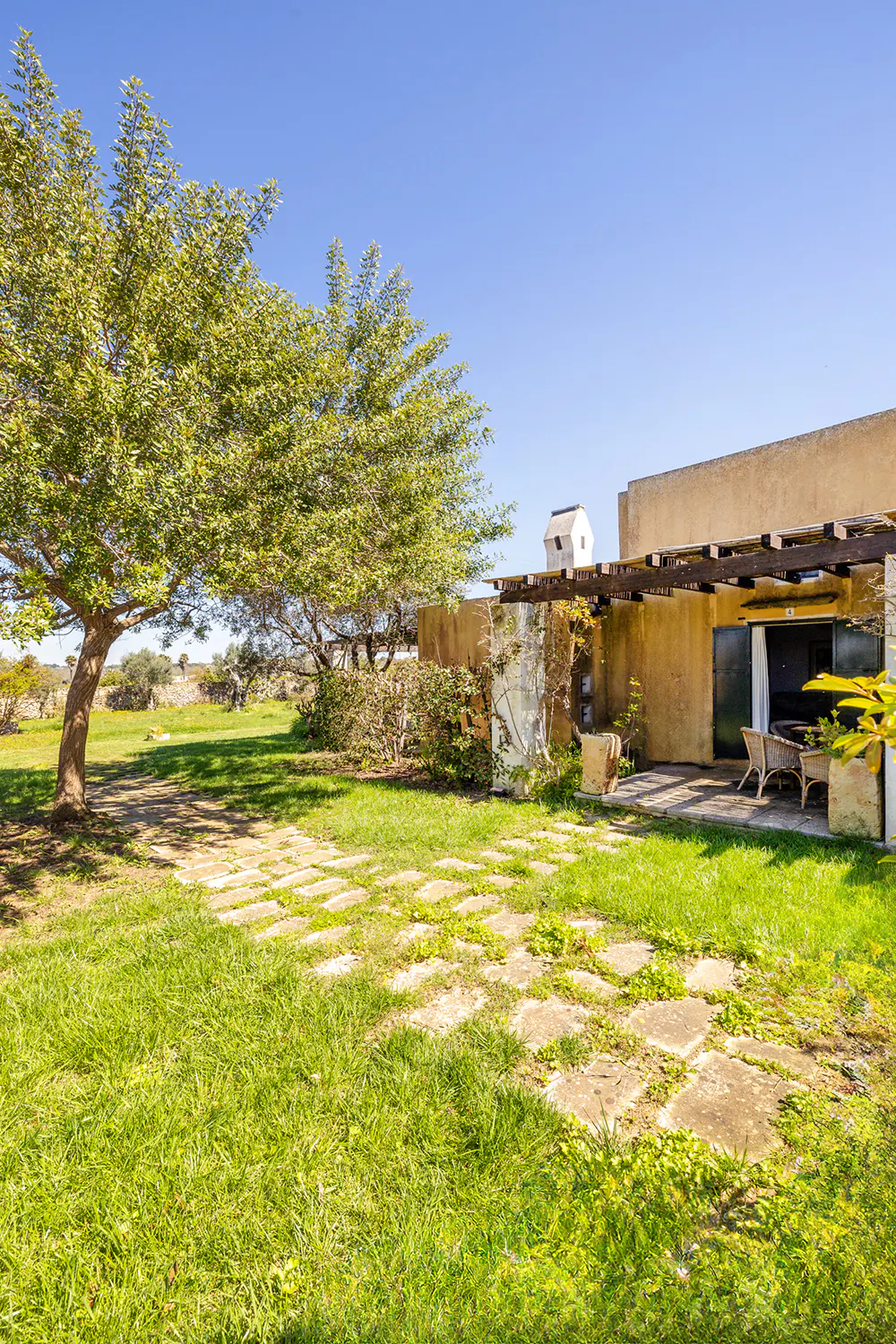 Exterior view of a tan house with a patio, chairs, and a large tree in the yard under a blue sky.