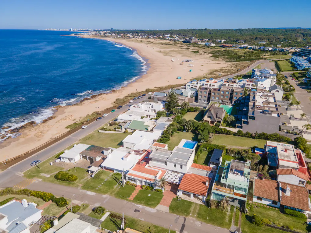 Aerial view of a coastal neighborhood with houses, a sandy beach, and the blue ocean under a clear sky.