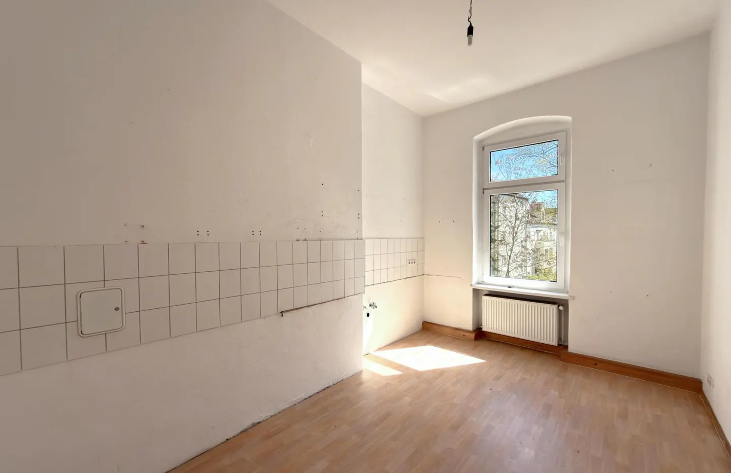 Empty kitchen with white walls, light wood floor, and a window showing a building and trees. White square tiles line the lower wall.