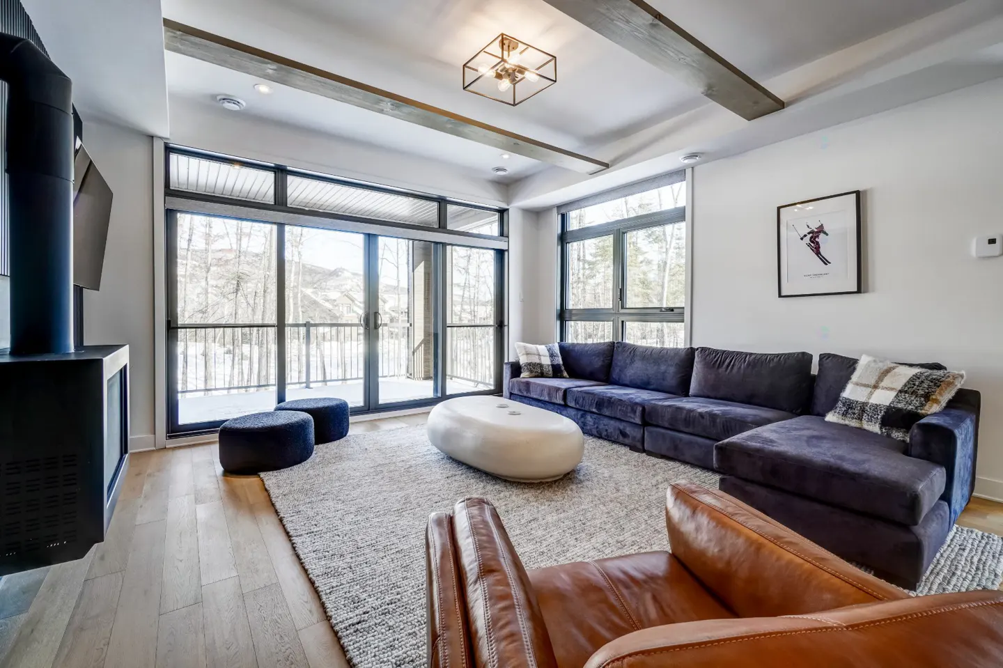 Living room with a blue sectional sofa, brown leather chair, white coffee table, and a fireplace. Large windows overlook a snowy landscape.