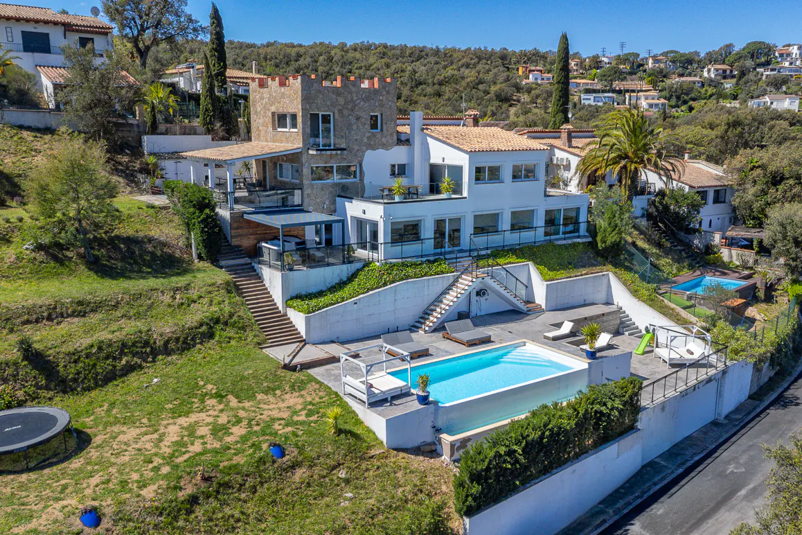 Aerial view of a modern white house with a pool, stone tower, and multiple levels on a green hillside.