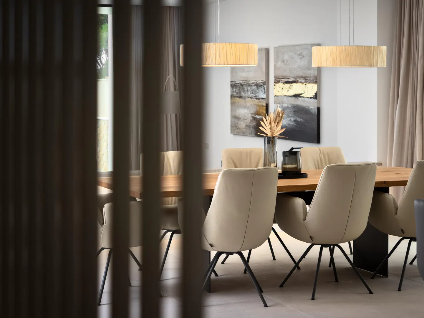 Dining room with a wood table, beige chairs, and modern light fixtures. Abstract art hangs on the wall. A vase with dried palms sits on the table.