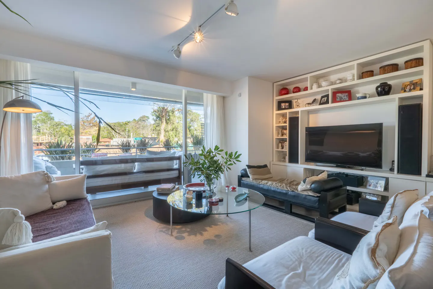 Bright living room with white sofas, a glass coffee table, and a large window overlooking a green landscape. A built-in shelf unit houses a TV and decorative items.