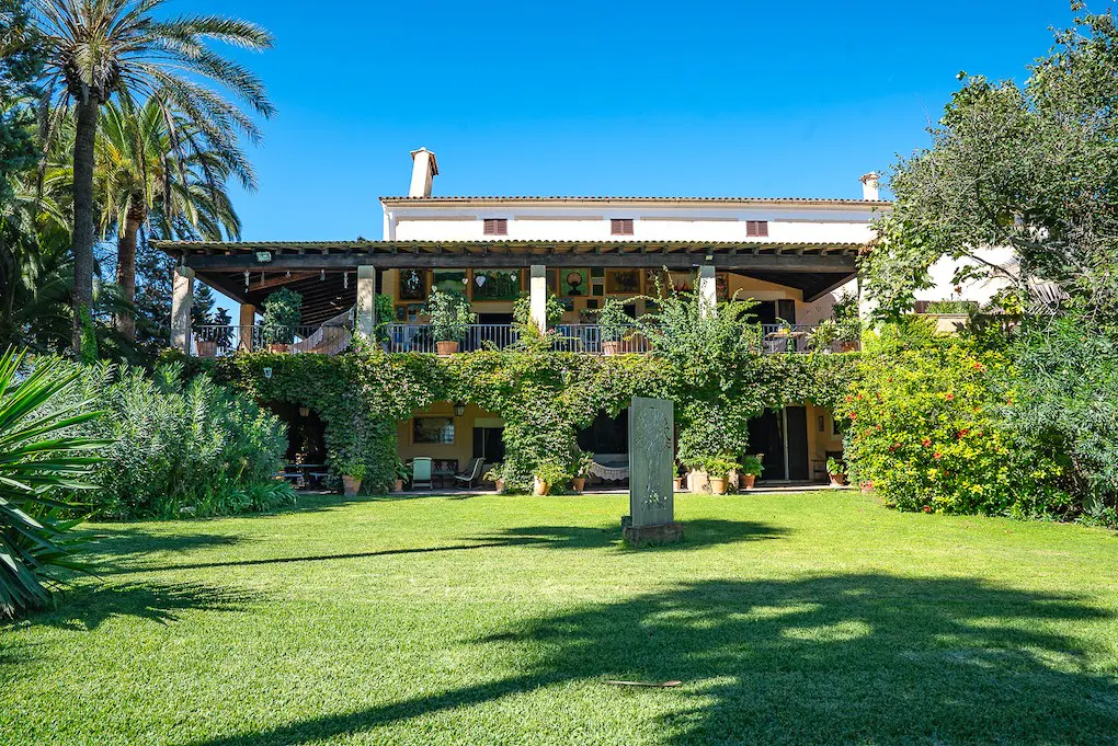 Two-story house with a large, covered balcony, lush green lawn, and palm trees under a clear blue sky.