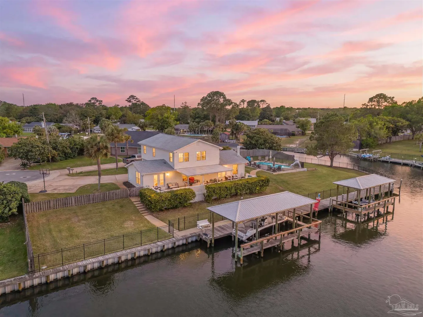 Aerial view of a two-story white house with a dock and boat lift on a canal at sunset.