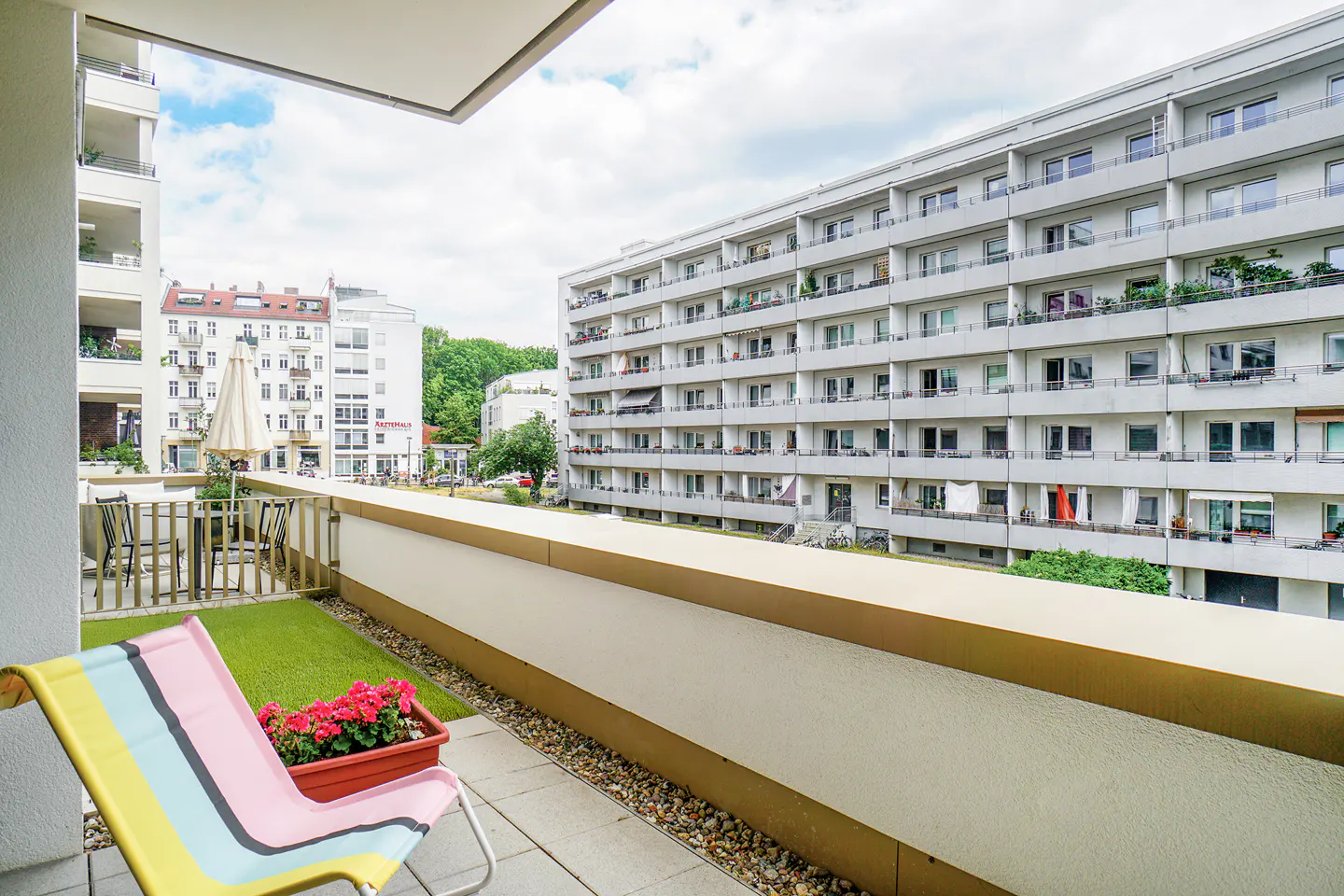 Balcony view with a colorful striped lounge chair, red flowers, and a table with chairs. Buildings are visible in the background.