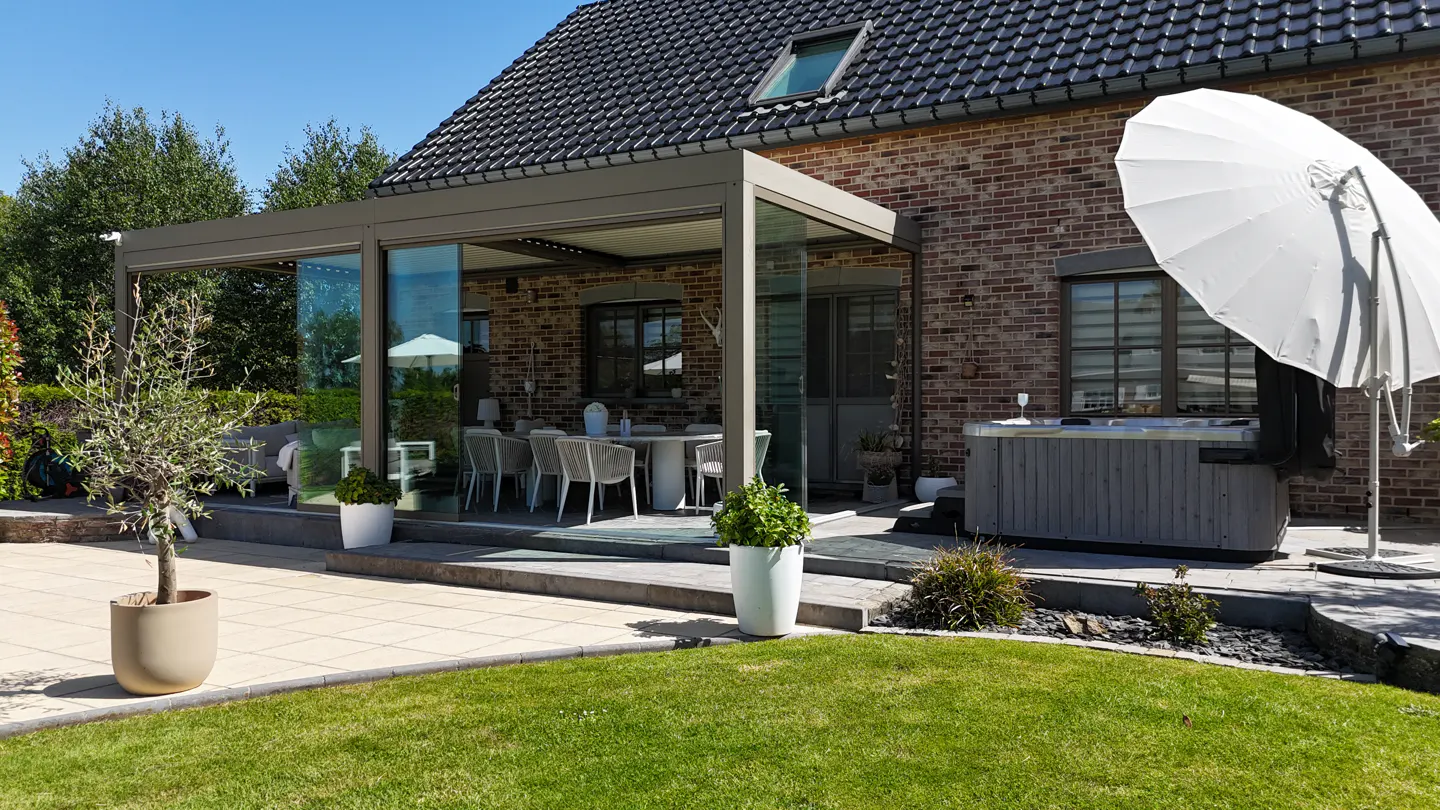 A brick house with a glass-enclosed patio, outdoor dining set, hot tub, and white umbrella on a sunny day.