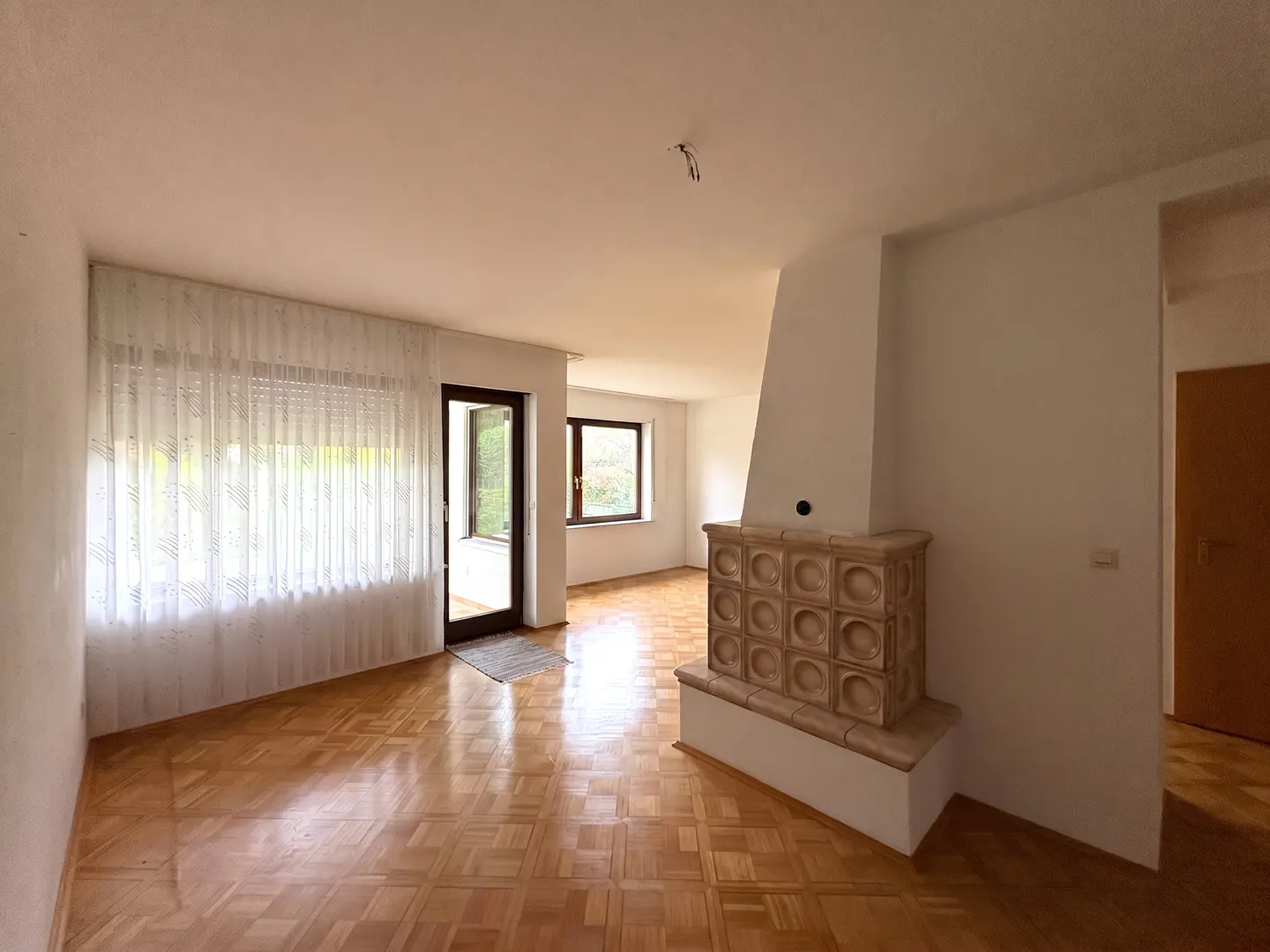 Bright, empty living room with parquet floors, white walls, and a tiled fireplace. A window with sheer curtains and a door to the outside are visible.