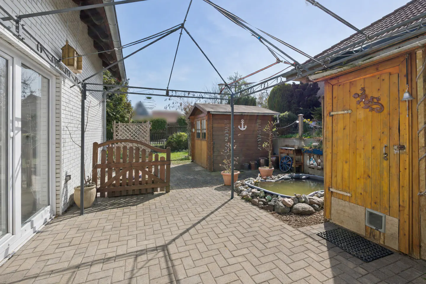 Backyard patio with brick pavers, metal pergola, wooden sheds, and a small pond with rock border.