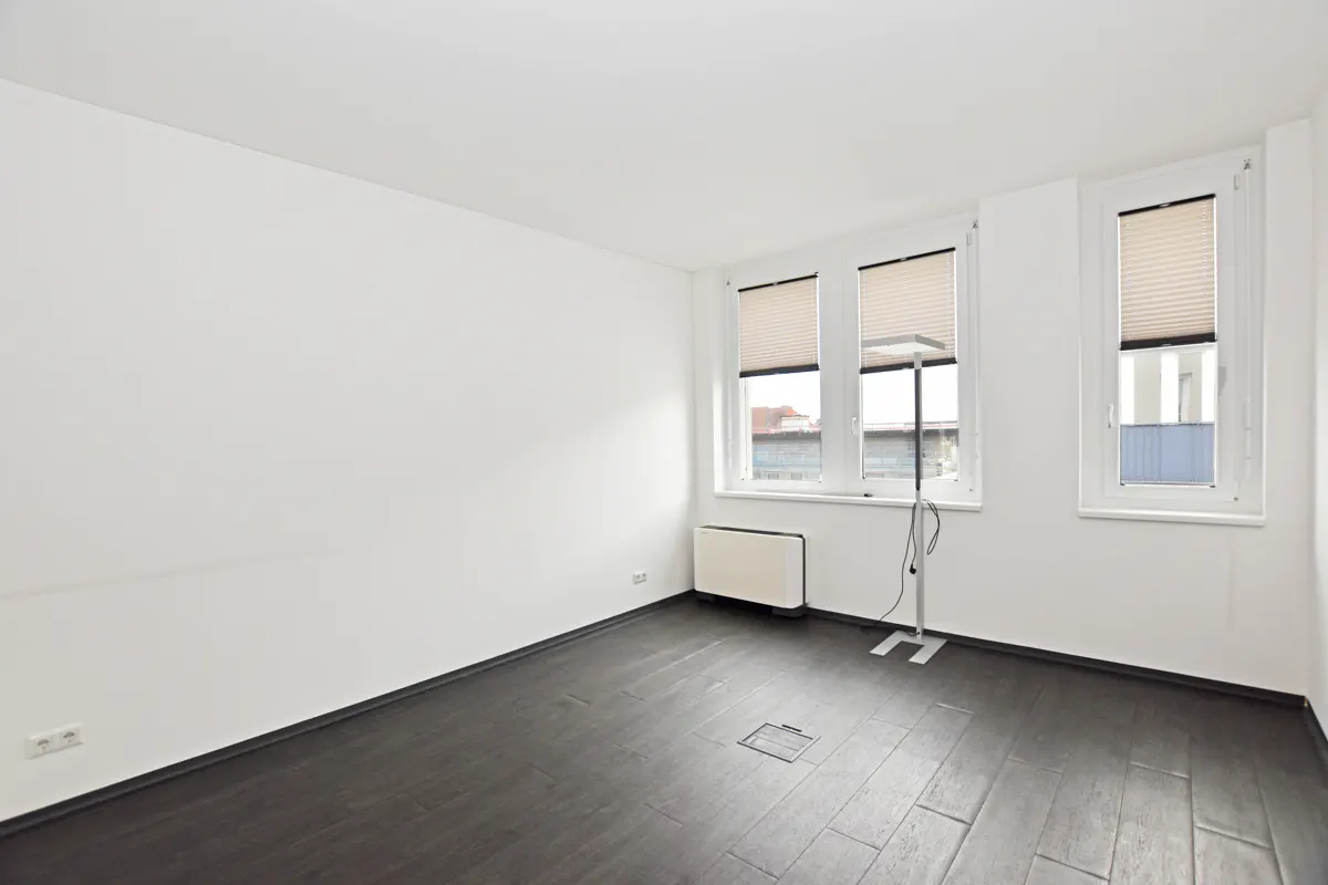 Bright, empty room with white walls, dark wood floors, and three windows with closed blinds. A white radiator sits under the windows.
