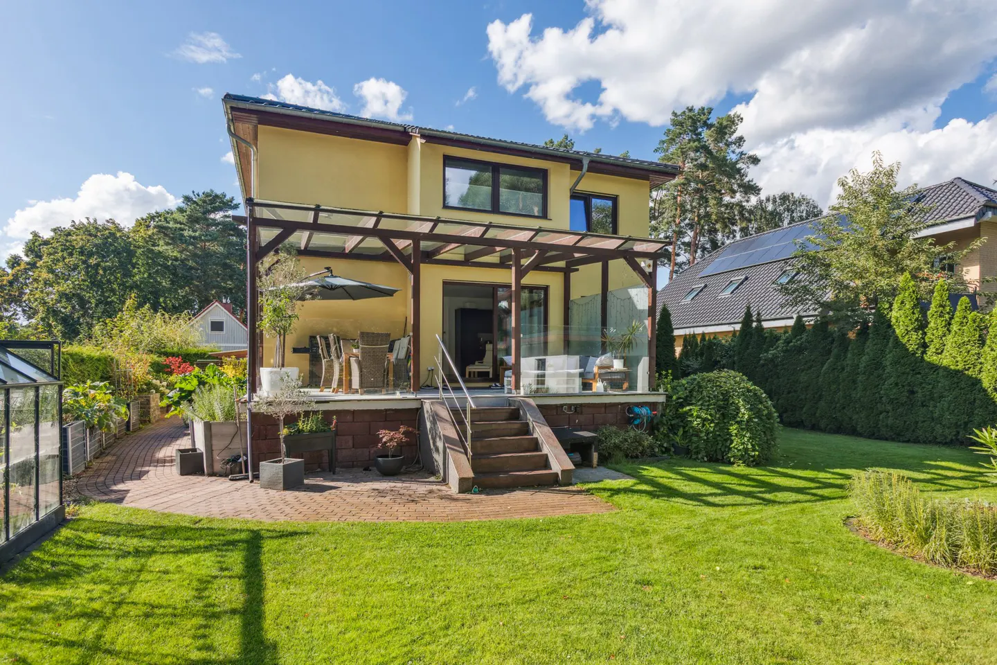 Backyard view of a two-story yellow house with a wooden pergola over a patio with outdoor furniture and a green lawn.