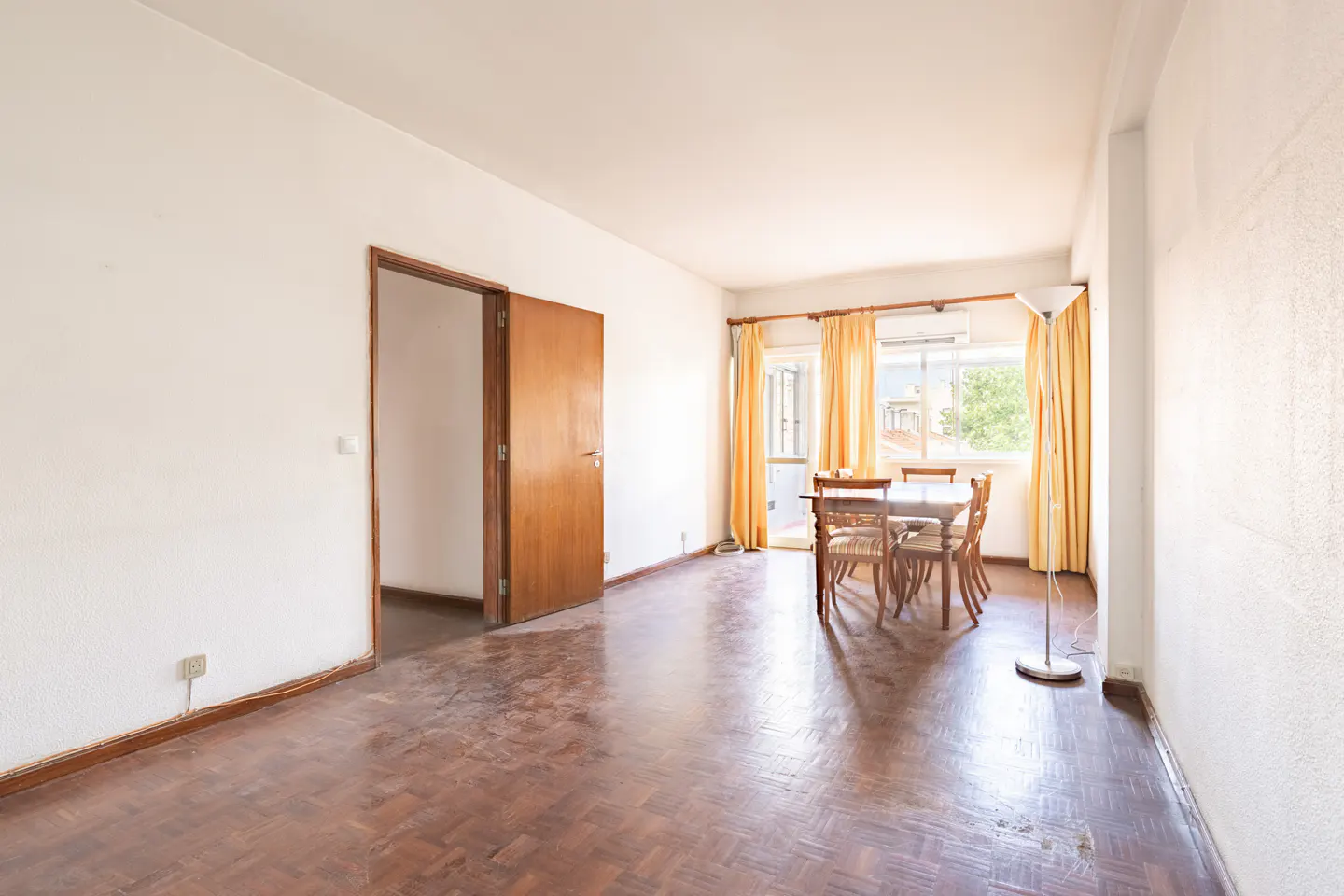 A bright, empty room with a dining table, wooden floors, and yellow curtains.