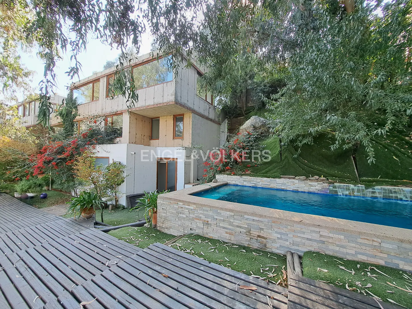 Exterior view of a modern, multi-story home with a pool, wooden deck, and lush greenery. The Engel & Völkers logo is visible.