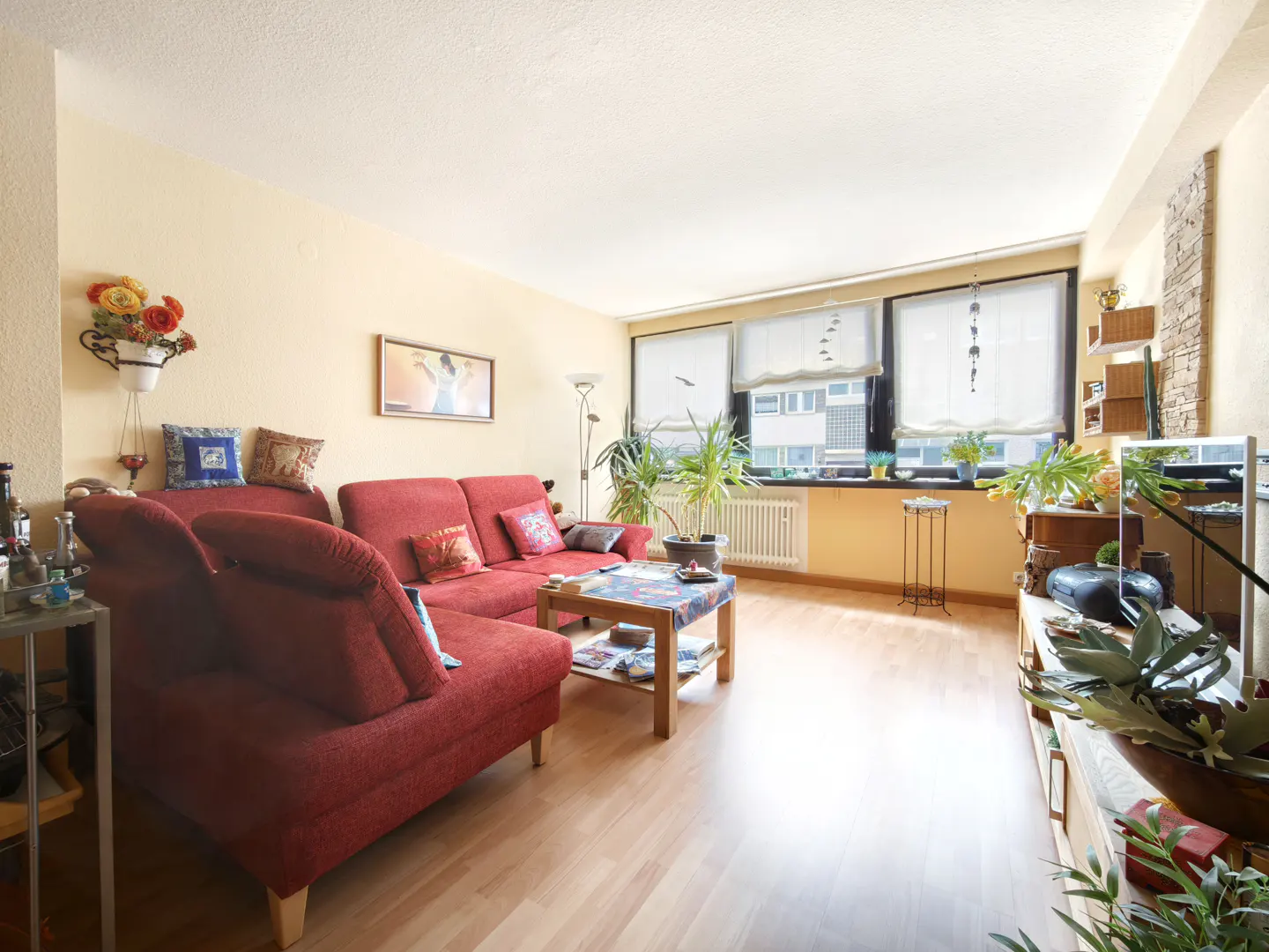 A bright living room with a red sectional sofa, wood floors, and large windows. Plants and decor add a homey touch.