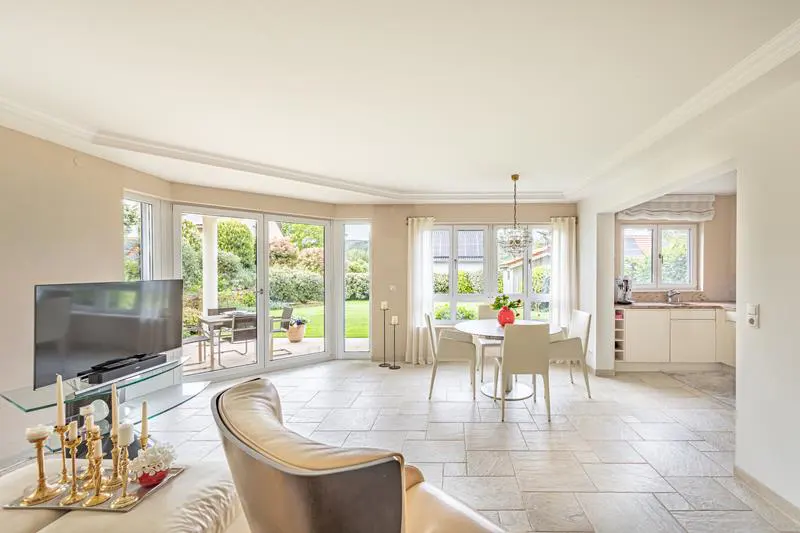 Bright, open-plan living space with tile flooring, a dining area, and a view of a lush green garden through large windows. A TV and seating area are visible.