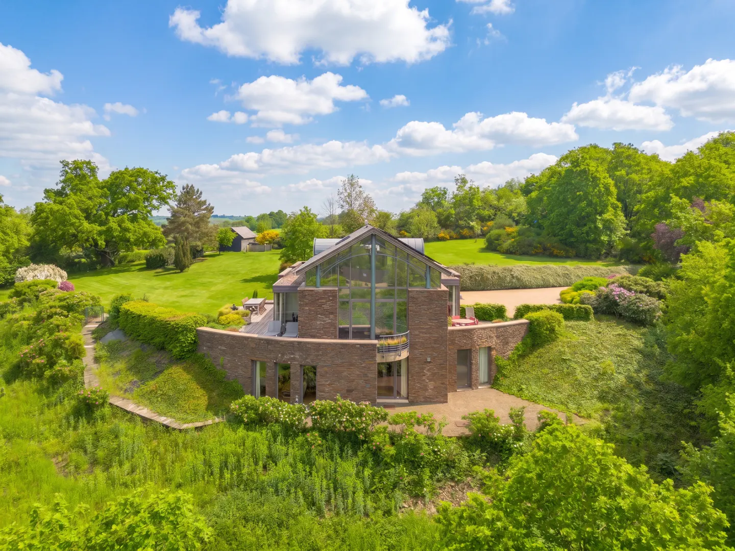 Aerial view of a modern brown brick house with a glass facade, surrounded by green trees and a lawn under a blue sky.