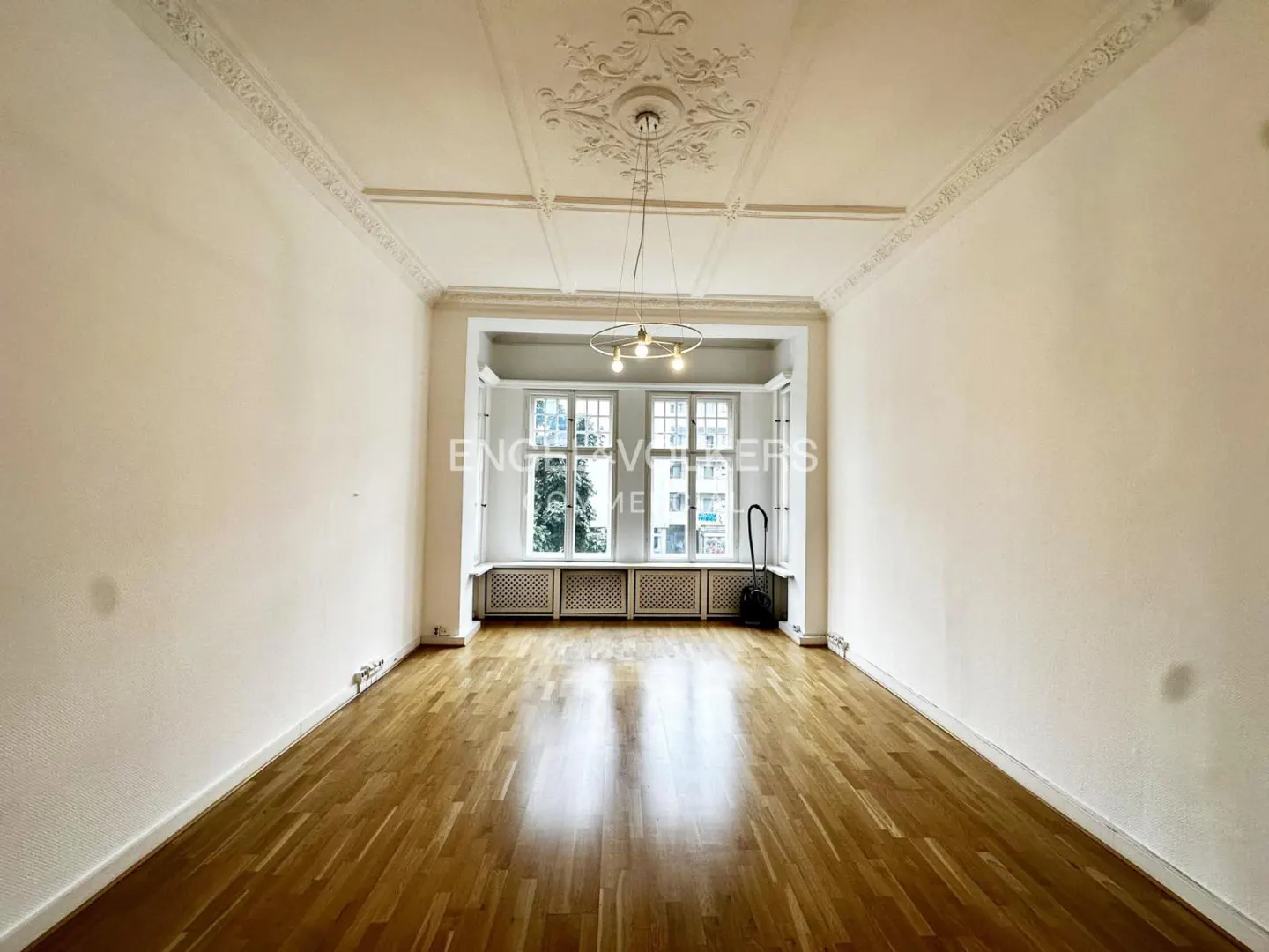 Empty room with hardwood floors, white walls, and a decorative ceiling. Windows overlook a city street. A modern chandelier hangs from the ceiling.