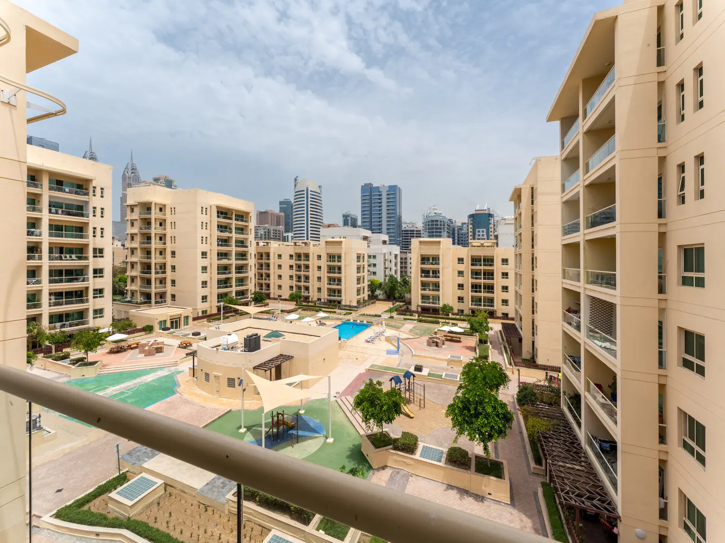View from a balcony overlooking a courtyard with a pool, playground, and beige apartment buildings. City skyline in the background.