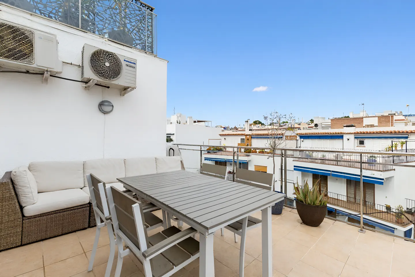 Rooftop patio with a gray table, chairs, and a wicker sofa. White buildings and a blue sky are in the background.